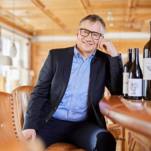 Hotel Sonnenhof Mann mit Brille in Anzug sitzt an Bar mit Weinflaschen im hellen Raum.