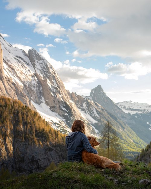 Hotel Sonnenhof Frau sitzt mit Hund auf Bergwiese mit Blick auf schneebedeckte Berge