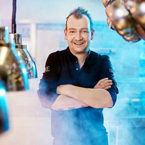 Hotel Sonnenhof Lächelnder Koch mit verschränkten Armen in einer dampfigen Küche