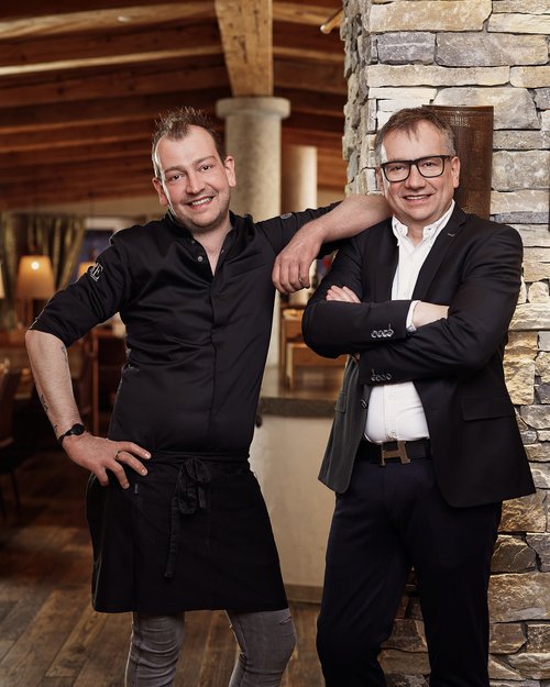 Hotel Sonnenhof Zwei lächelnde Männer, einer in Kochkleidung, der andere im Anzug, lehnen an einer Steinwand