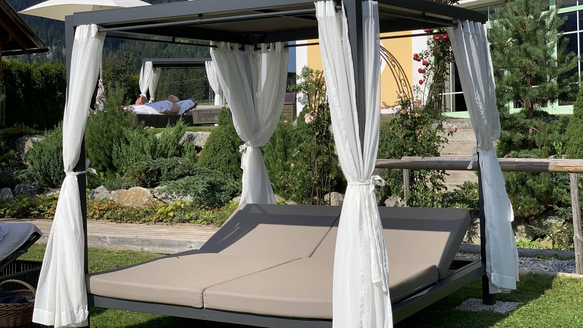 Hotel Sonnenhof Outdoor-Lounge-Doppelbett mit Vorhängen vor Blumen und Bergkulisse