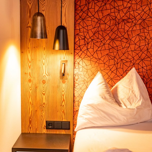 Hotel Sonnenhof Betthaupt mit rotem Muster, Nachttisch und zwei Hängelampen an Holzwand