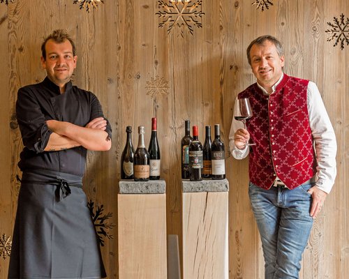 Hotel Sonnenhof Zwei Männer, einer Koch in Schwarz, der andere mit Glas Wein vor Holzwand mit Weinflaschen