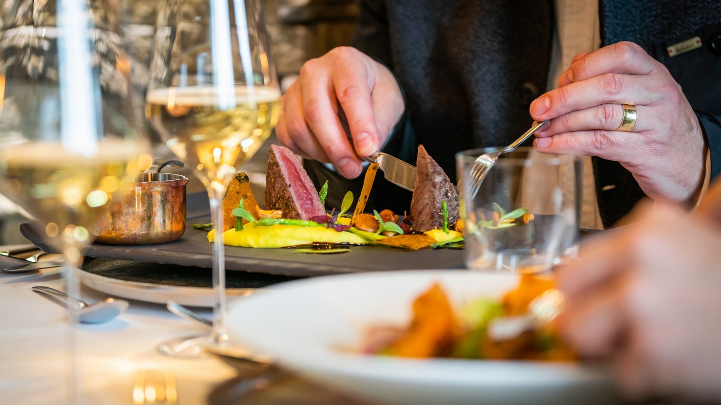 Hotel Sonnenhof Person schneidet Fleisch bei gehobenem Essen mit Weingläsern auf Tisch