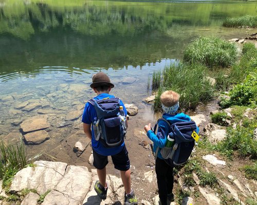 Hotel Sonnenhof Zwei Kinder mit Rucksäcken am Ufer eines klaren Bergsees