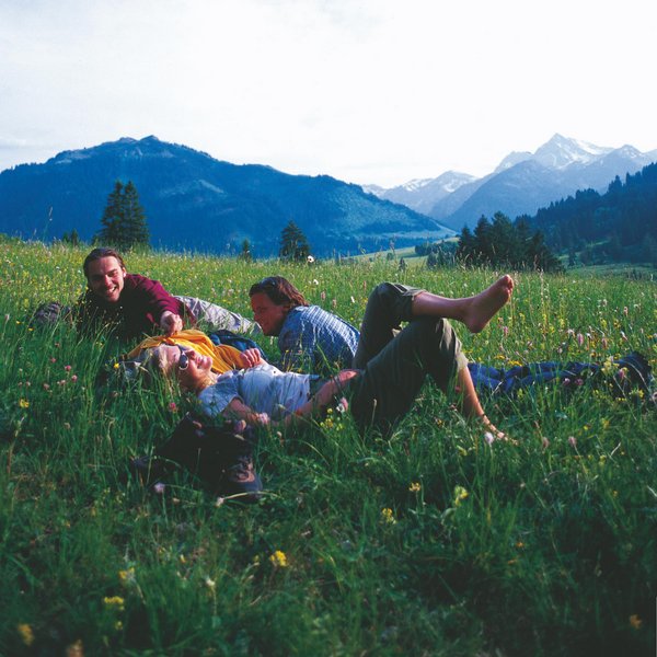 Hotel Sonnenhof Drei Menschen liegen entspannt auf einer Blumenwiese mit Bergen im Hintergrund
